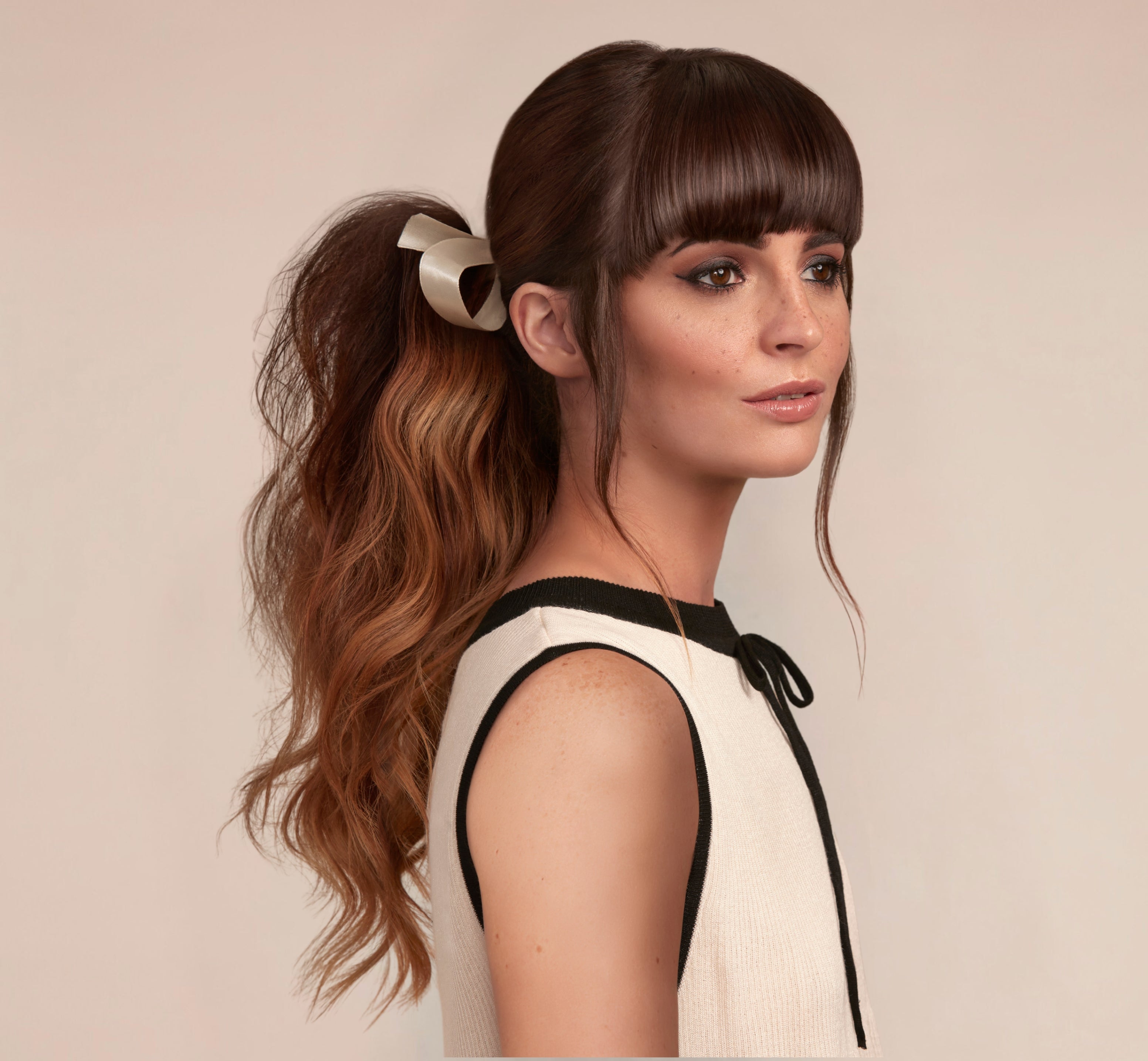 Woman with brunette styled hair in a wavy textured ponytail and a sleek glossy straight fringe wearing a white top with black accents.