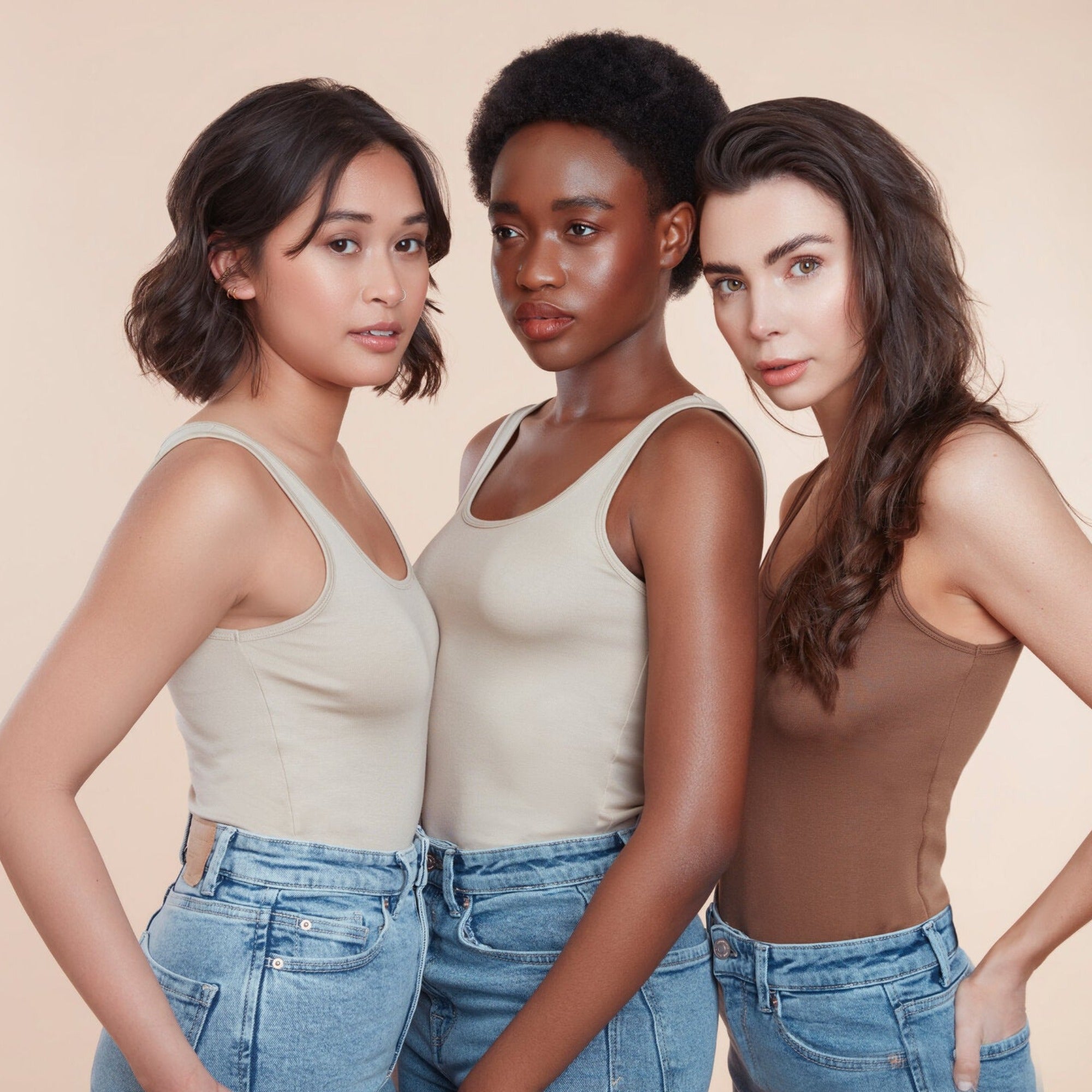 Three female models with moo and yoo products in their hair all wearing jeans and neutral coloured vest tops. They are looking directly at the camera. All have glowing skin and healthy shiny hair. One girl has bob length wavy hair, One girl has black natural textured hair and the other girl has long brown hair. 