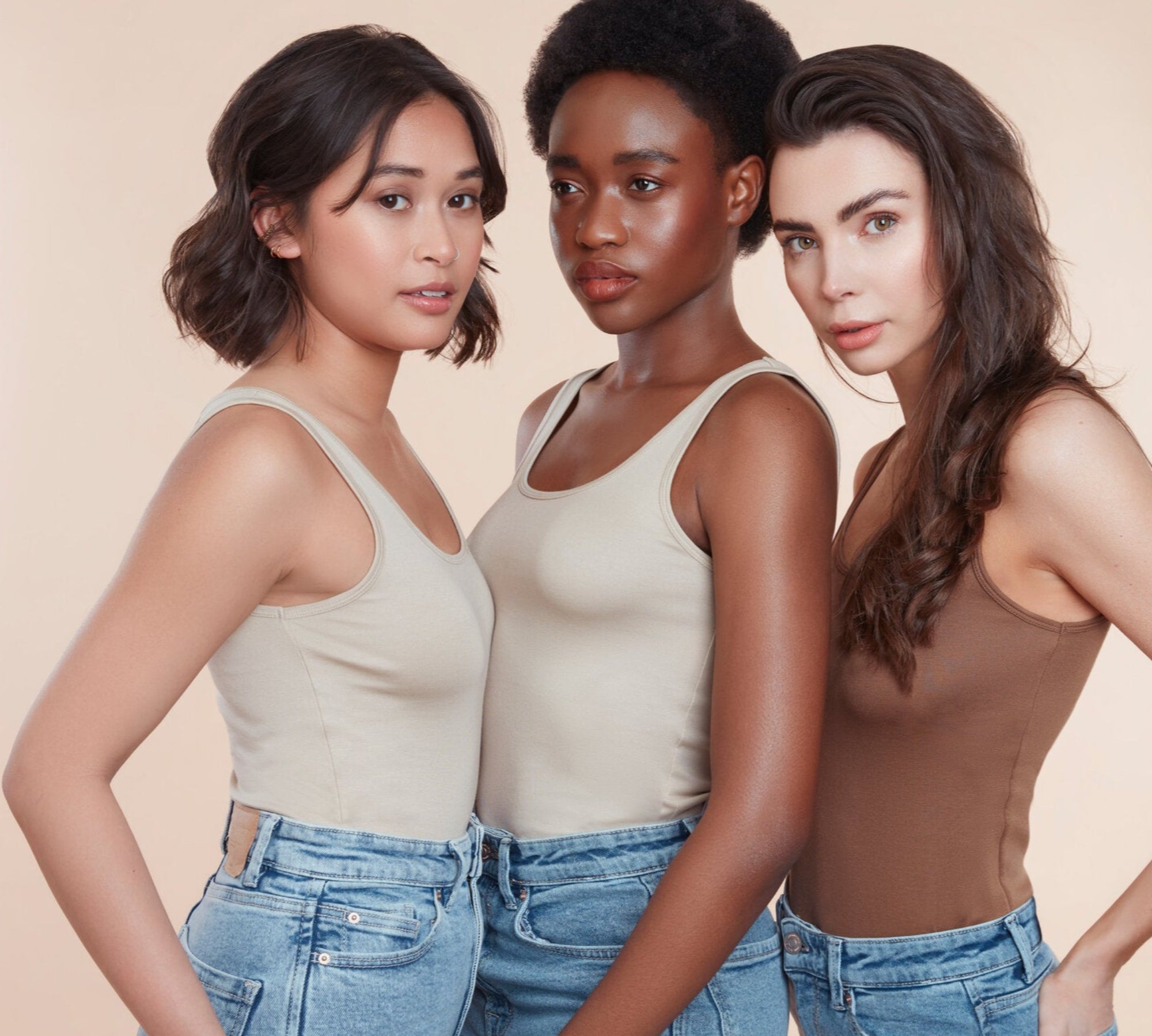 Three female models with moo and yoo products in their hair all wearing jeans and neutral coloured vest tops. They are looking directly at the camera. All have glowing skin and healthy shiny hair. One girl with bob length wavy hair, One with black natural textured hair and one with long brown hair. Model with long flowing brunette hair. She has super shiny hair with lots of volume.