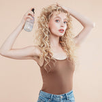 Female model with flowing blond curly hair. She is holding Moo and Yoo Sea Salt Spray in her right hand and spraying it into her hair, she is touching her hair with her left hand.  She is wearing a simple natural coloured vest top and her make up is very natural.