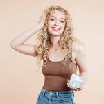 Smiling female model holding Moo and Yoo Miracle Curl Cream in one hand with her other hand in her curls. She has blond curls and is wearing a neutral coloured vest top.