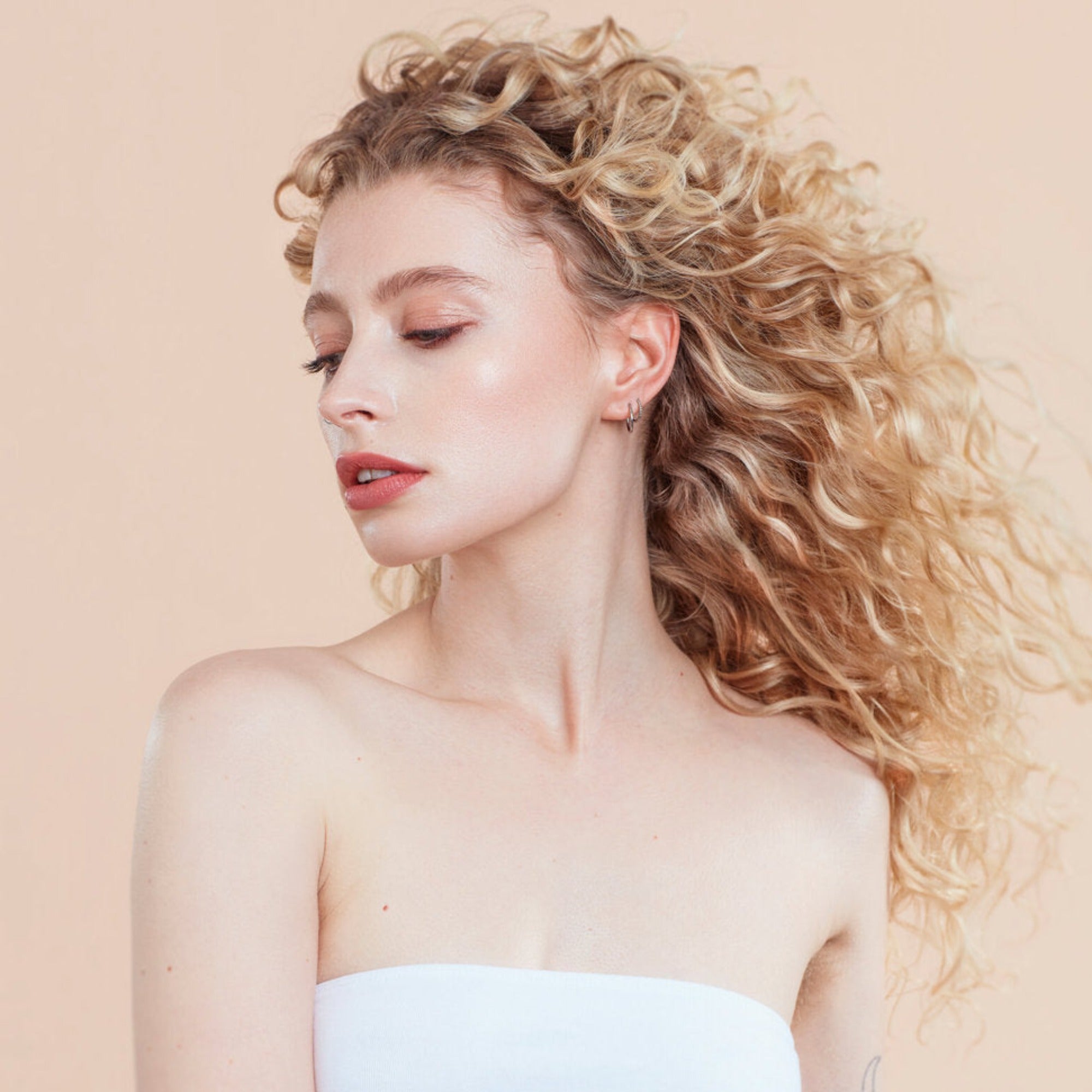 Female model with blond curly hair with moo and yoo conditioner followed by curl cream in her hair.  She is looking to her right and her hair is flowing. She is wearing a white bandeau. 
