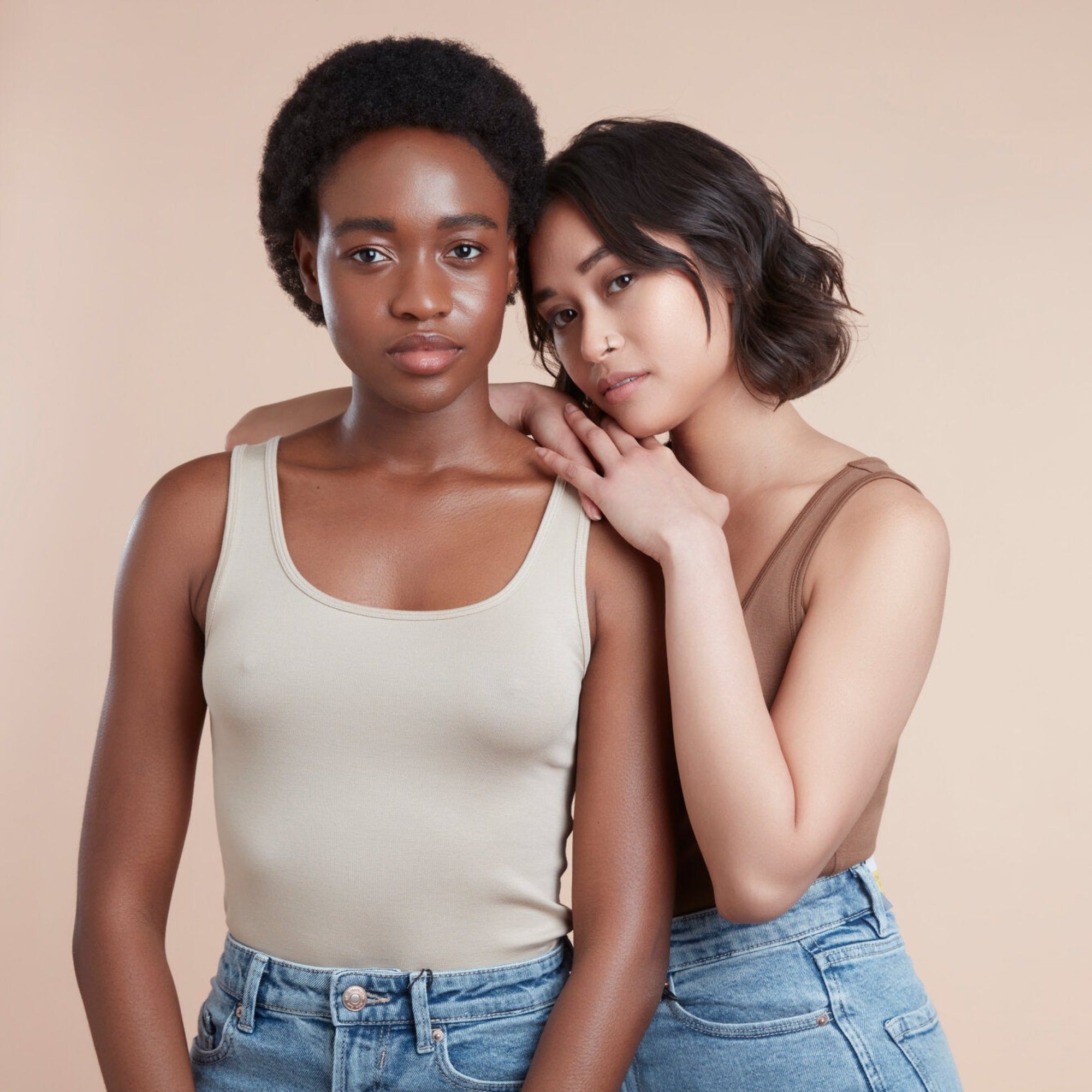 Two female models with moo and yoo products in their hair, both wearing jeans and neutral coloured vest tops. They are looking directly at the camera and one is leaning on the other. They have glowing skin and healthy shiny hair. One has bob length wavy hair, One has black natural textured hair.