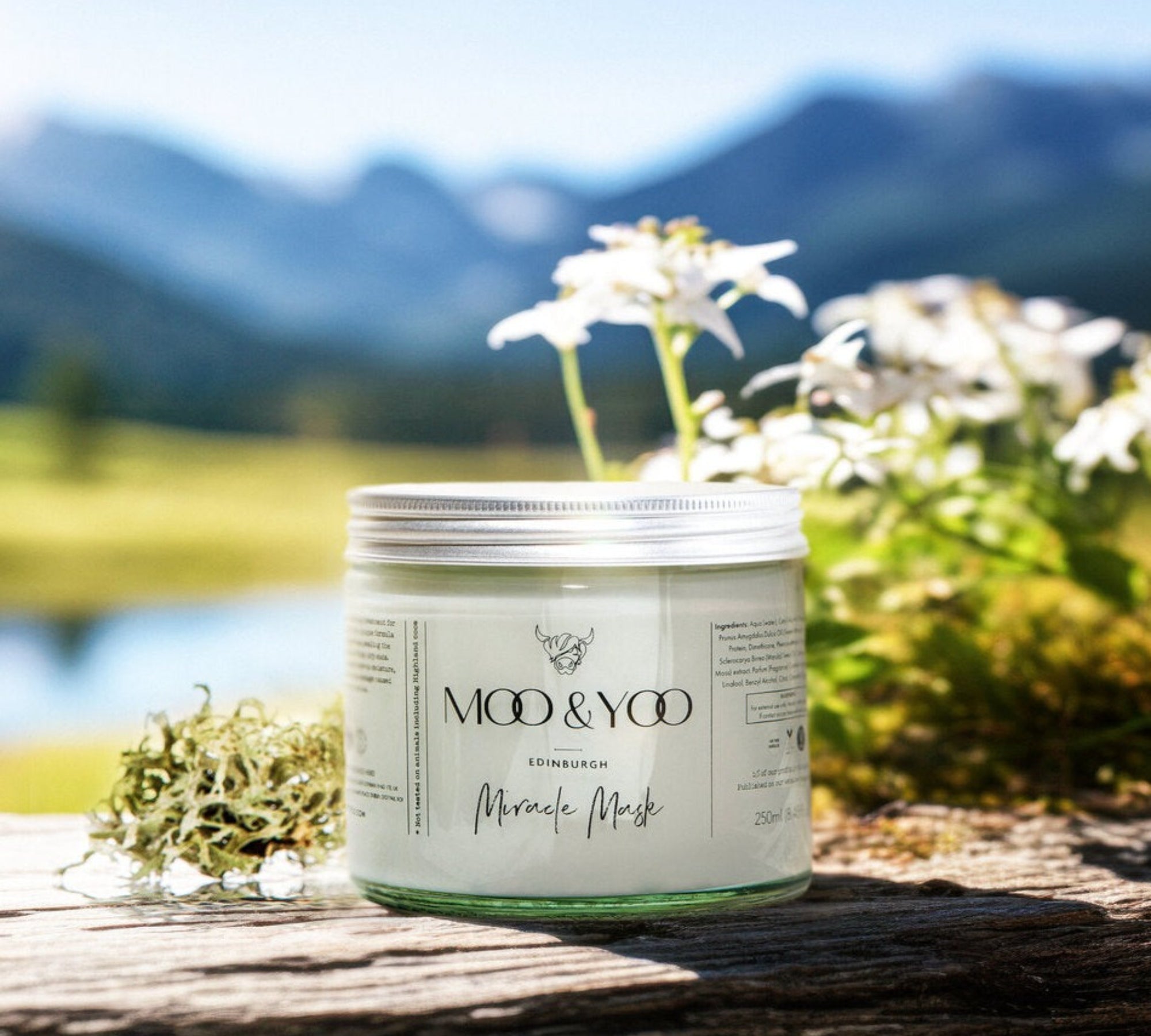 A glass jar of Moo and Yoo Miracle Mask with an aluminium lid. It is placed on a rustic wooden bench overlooking a Scottish Loch and hills.  It is a spring day and there are white spring flowers behind it.