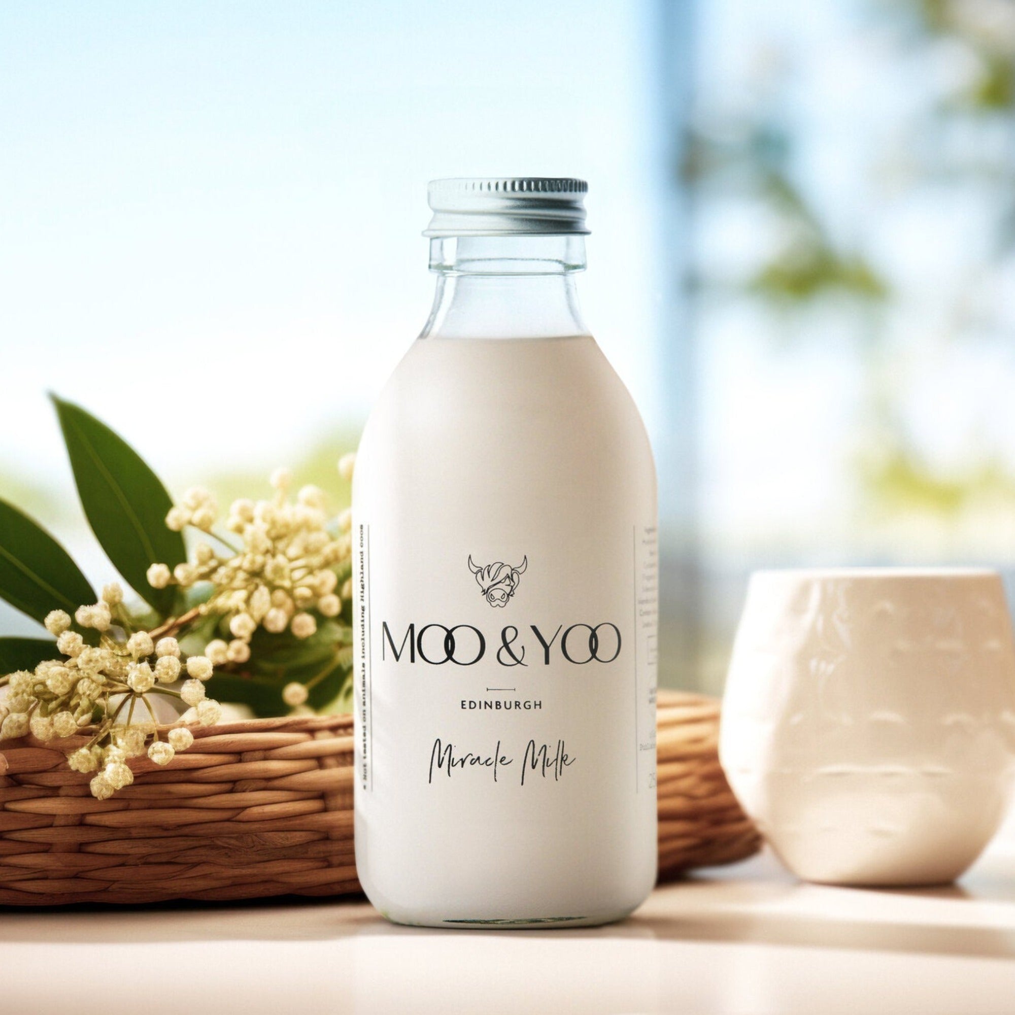 A glass bottle of Moo and Yoo Miracle Milk with an aluminium lid. It is placed on a white table with a basket with some small white flowers to one side and a small white plant pot to the other side.