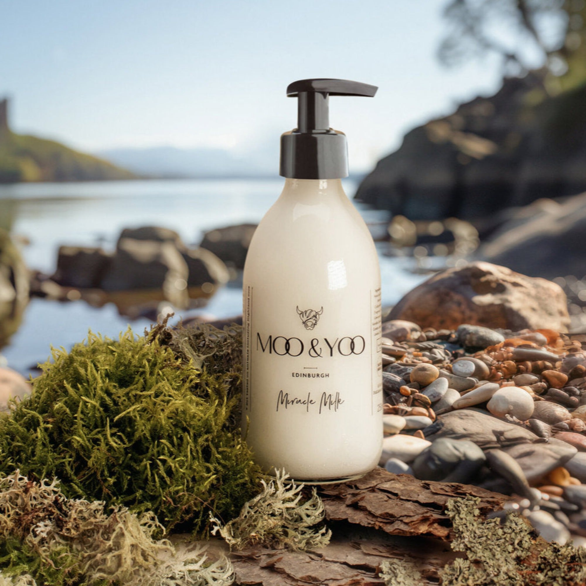 A glass bottle of Moo and Yoo Miracle Milk with a pump at the side of a Scottish Loch sitting on some pebbles and moss.