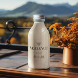 A glass bottle of Moo and Yoo Miracle Milk with an aluminium lid. It is placed on a teak bench overlooking the Scottish hills.  It is autumn and there is a plant pot with an autumn plant in and the leaves in the back ground are orange and yellow.