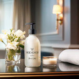 A glass bottle of Moo and Yoo Miracle Milk with a pump. It is placed on a table in a hotel suite. There is a towel and a candle to one side and a small vase of white flowers to the other side.  