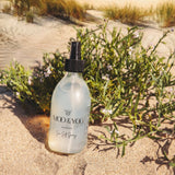 A glass bottle of Moo and Yoo Sea Salt Spray Milk sitting in the sand leaning on a small green shrub.