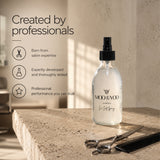 A clear glass spray bottle labeled 'MOD & YOOO Edinburgh Sea Salt Spray' stands on a stone surface in a warm, sunlit salon-like setting. Scissors and a comb rest nearby, with promotional text on the left reading 'Created by professionals.