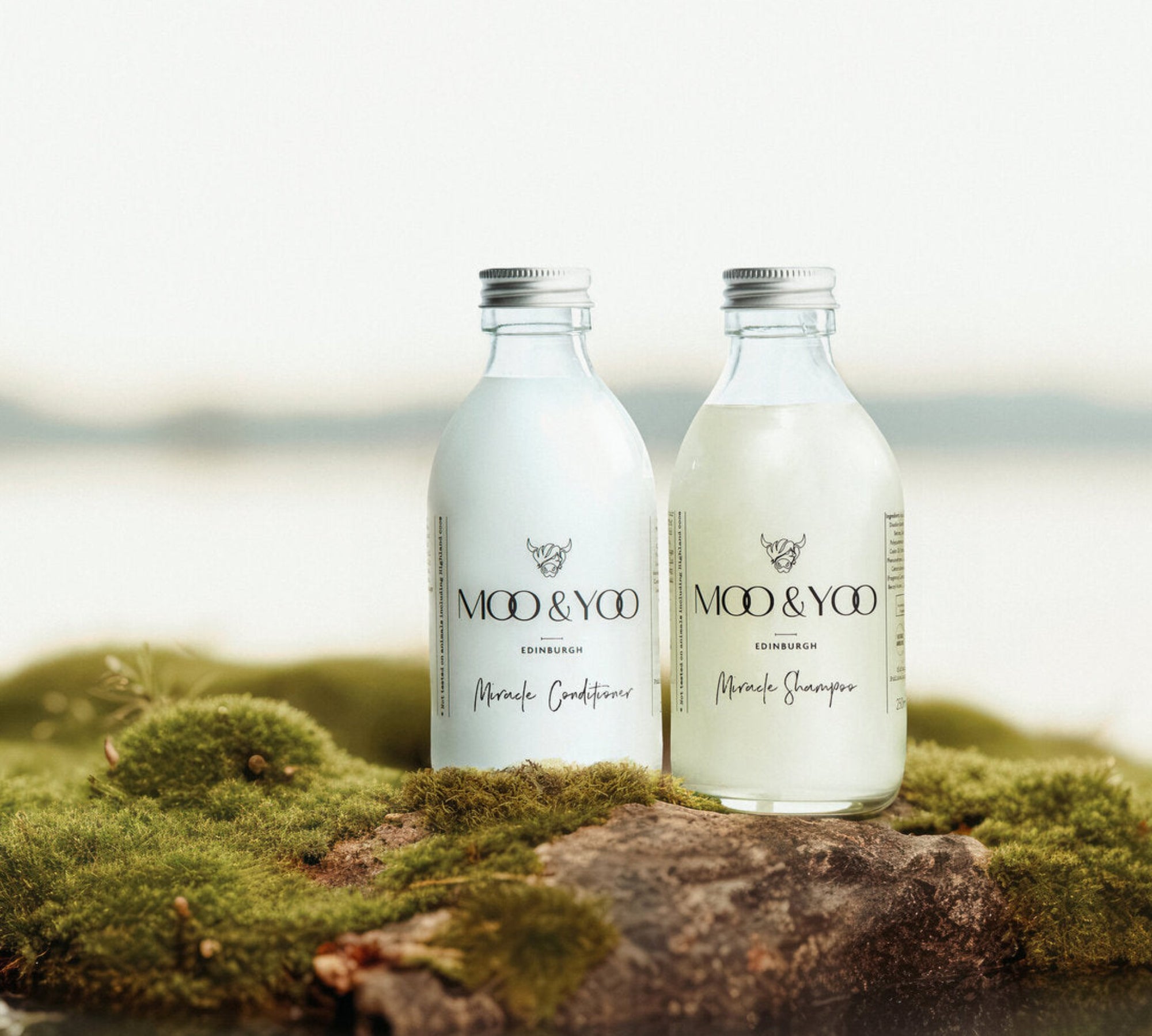 A glass bottle of Moo and Yoo Miracle Conditioner and a glass bottle of Moo and Yoo Miracle Shampoo sitting side by side with  aluminium lids. They are placed on a rock with moss in the edge of the water.
