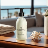 A close up image of a glass bottle of Moo and Yoo Miracle shampoo with a pump . It is placed on table in a hotel suite. There is a folded towel to one side and a plant to the other.  In the hazy background you can see a beach and the sea.