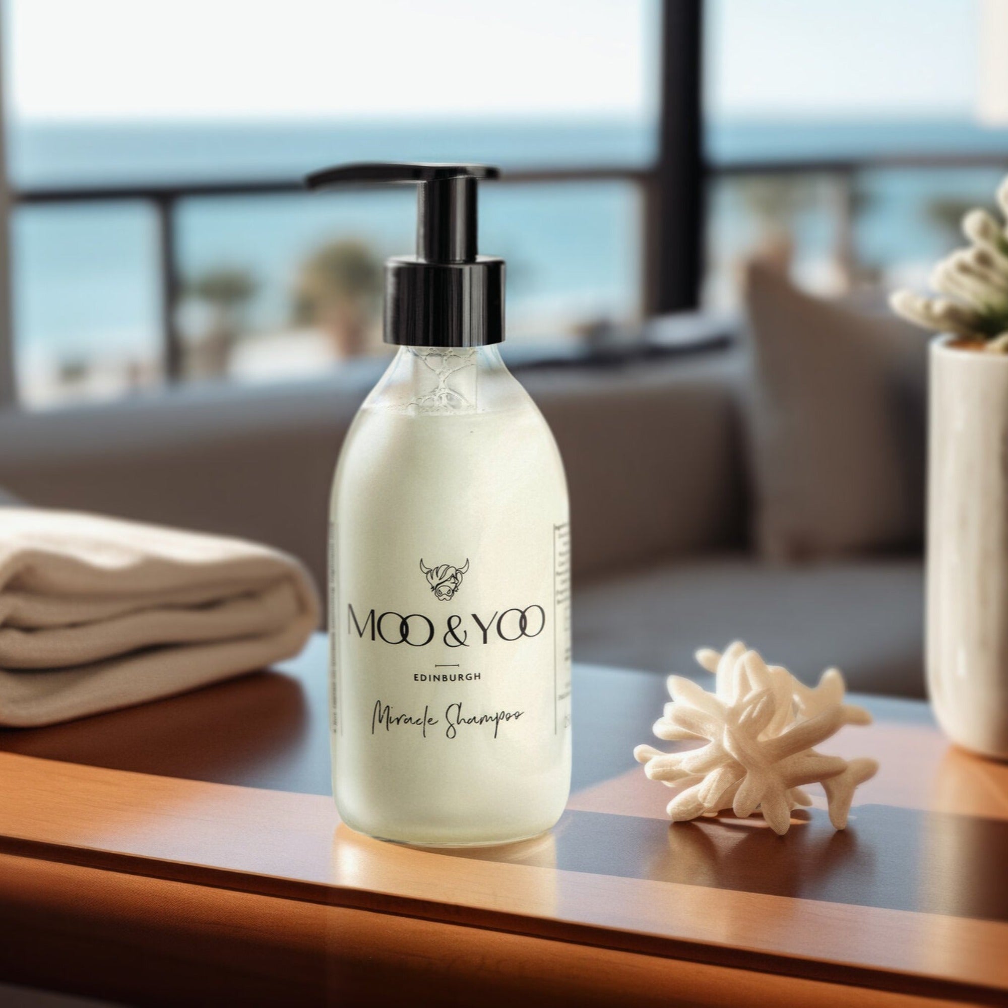 A close up image of a glass bottle of Moo and Yoo Miracle shampoo with a pump . It is placed on table in a hotel suite. There is a folded towel to one side and a plant to the other.  In the hazy background you can see a beach and the sea.