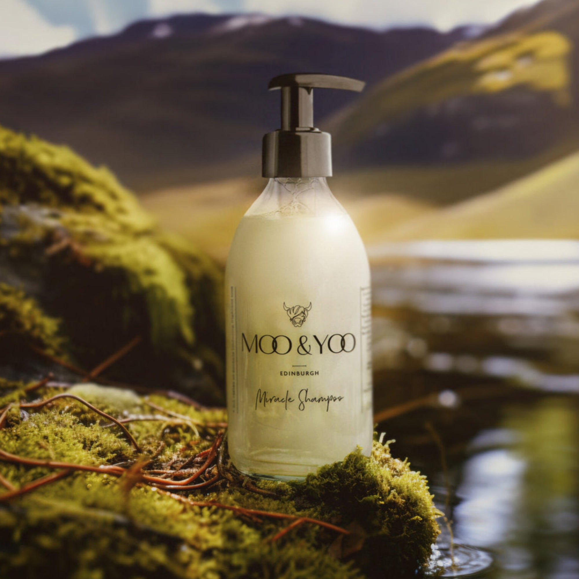 Our miracle shampoo in a glass bottle on a moss stone on a river bank. The river leads to the background of a valley in-between the Scottish mountains and lots of greenery