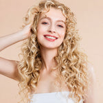 A smiling woman with long, voluminous curly blonde hair, wearing a white strapless top against a solid light beige background.