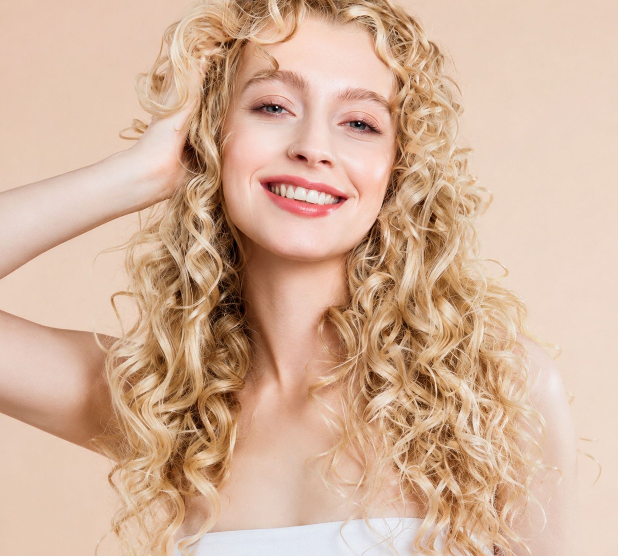 A smiling woman with long, voluminous curly blonde hair, wearing a white strapless top against a solid light beige background.