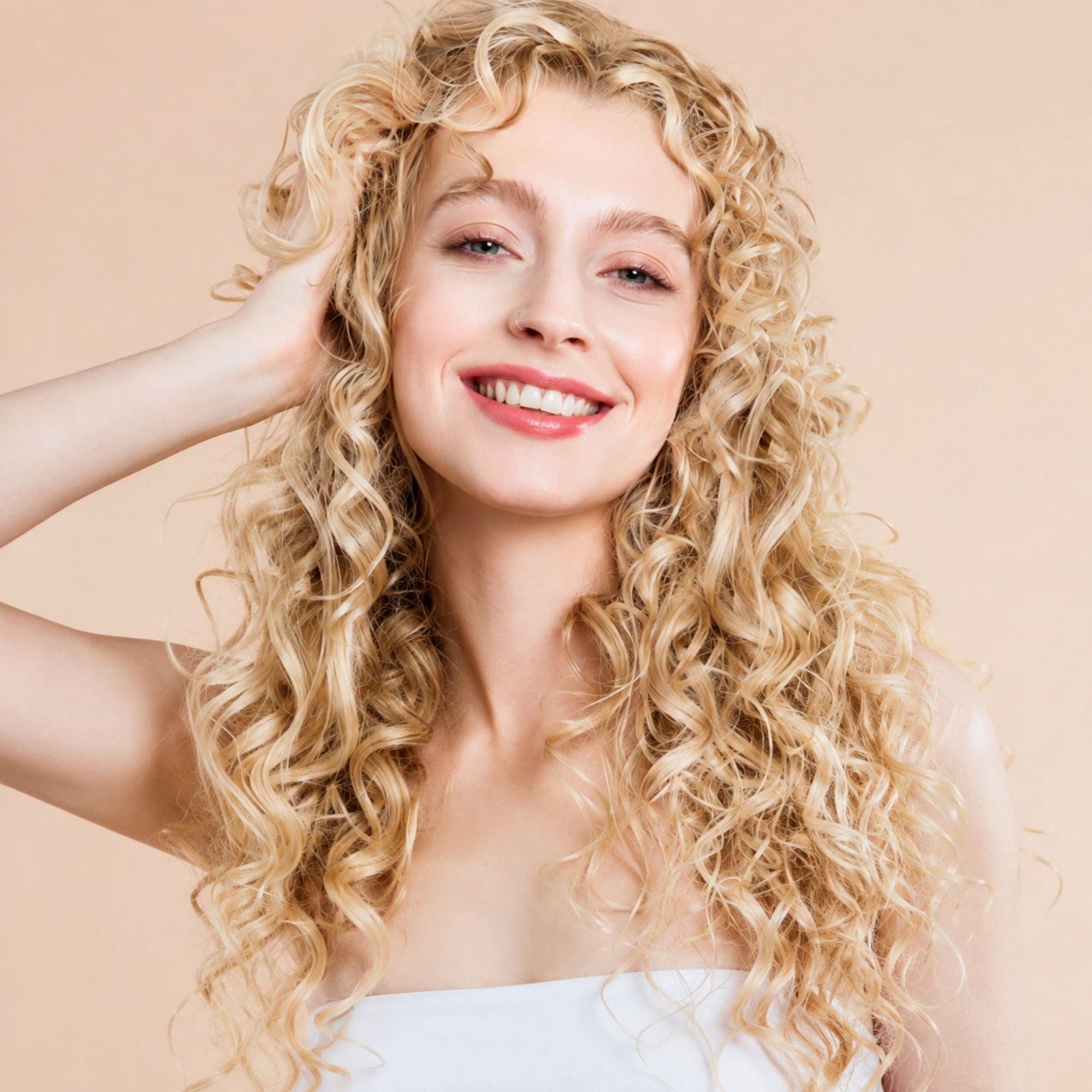 A smiling woman with long, voluminous curly blonde hair, wearing a white strapless top against a solid light beige background.