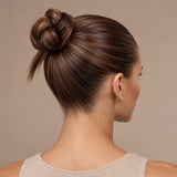 A woman shown from the back and side profile wearing a dark brown twisted bun hairstyle with subtle warm blonde highlights, against a solid beige background.