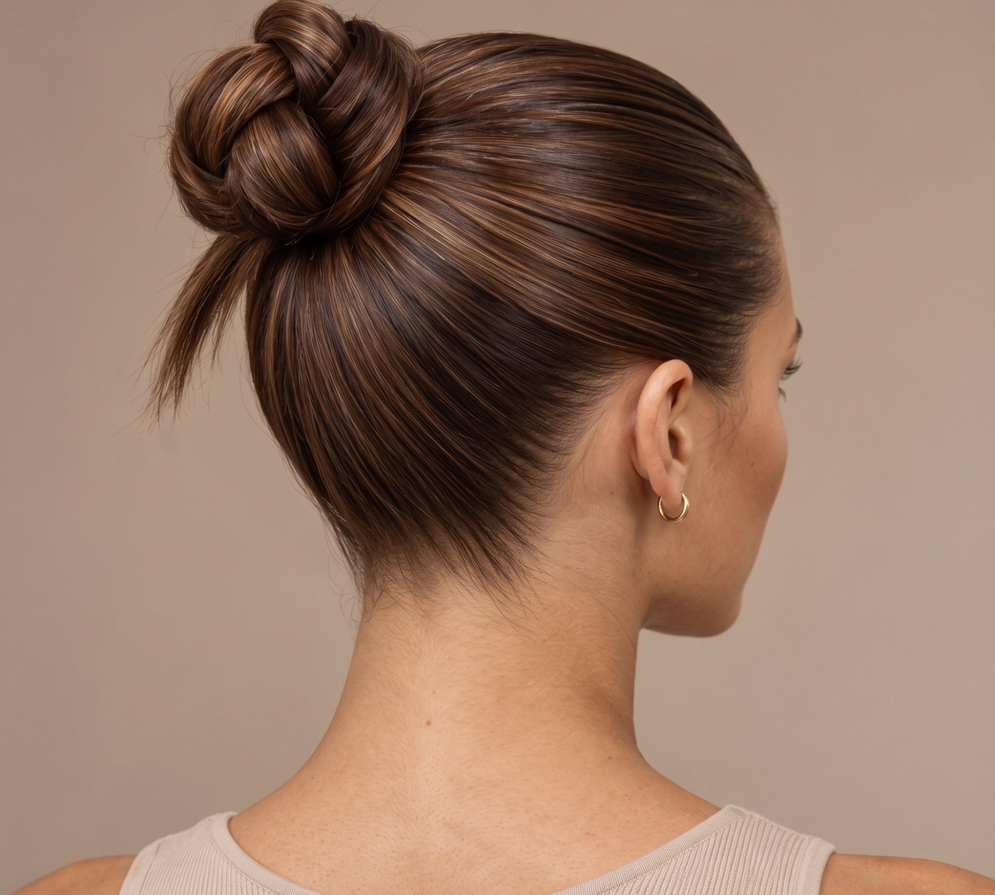 A woman shown from the back and side profile wearing a dark brown twisted bun hairstyle with subtle warm blonde highlights, against a solid beige background.