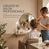 A salon scene with a stylist wearing a beige T-shirt and blue jeans assisting a client seated in a salon chair. A round wall mirror reflects the salon interior, and a bottle labeled Moo & yoo hair spray sits on the counter beside a brush and other salon accessories.