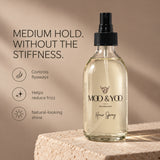 A clear glass moo & Yoo hair spray bottle with a black spray top stands on a stone surface against a warm beige background. The bottle label reads 'MOO & YOO EDINBURGH' and 'Hair Spray.