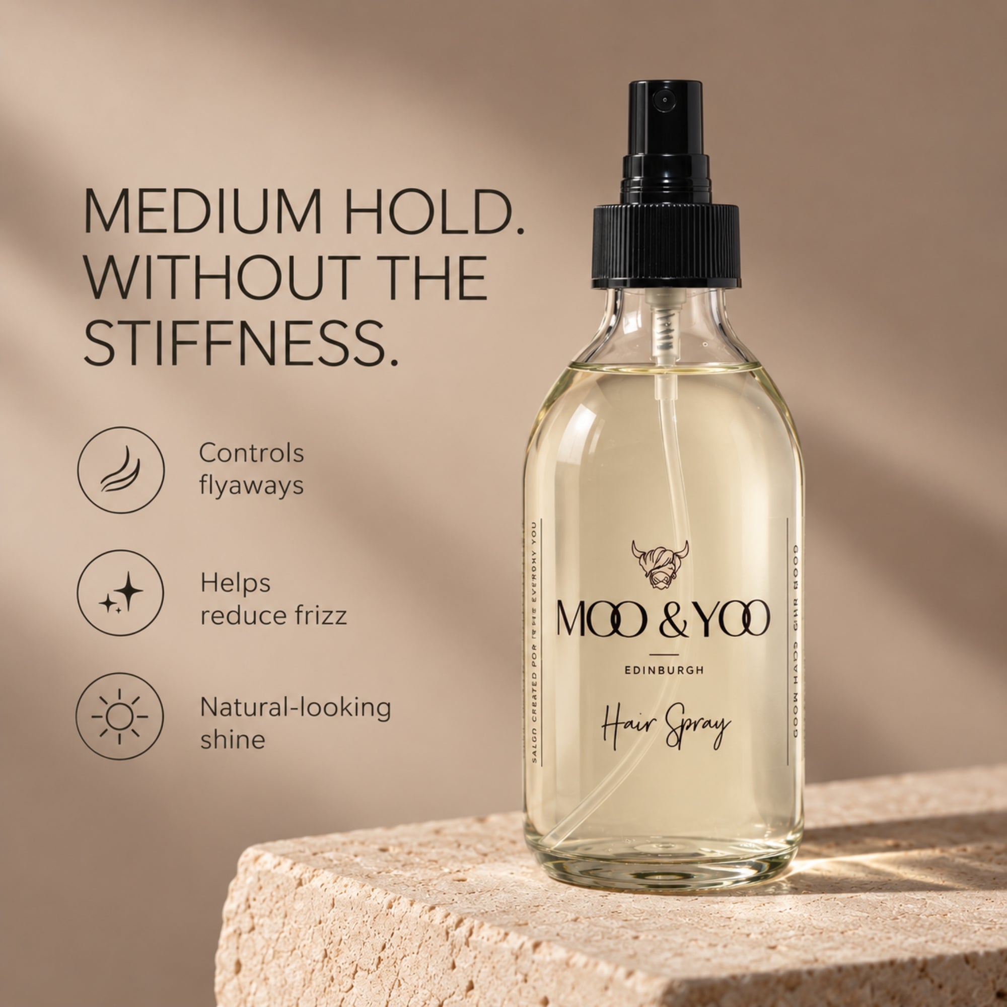 A clear glass moo & Yoo hair spray bottle with a black spray top stands on a stone surface against a warm beige background. The bottle label reads 'MOO & YOO EDINBURGH' and 'Hair Spray.