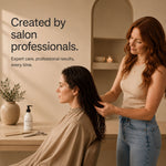 A lifestyle scene in a warm, minimalist salon setting showing a hairstylist with long red hair adding some moo and yoo miracle milk into the the wet dark hair of a seated client. The background features a warm beige wall with an arched shelf, a small potted plant, and a bottle of hair product on the counter.