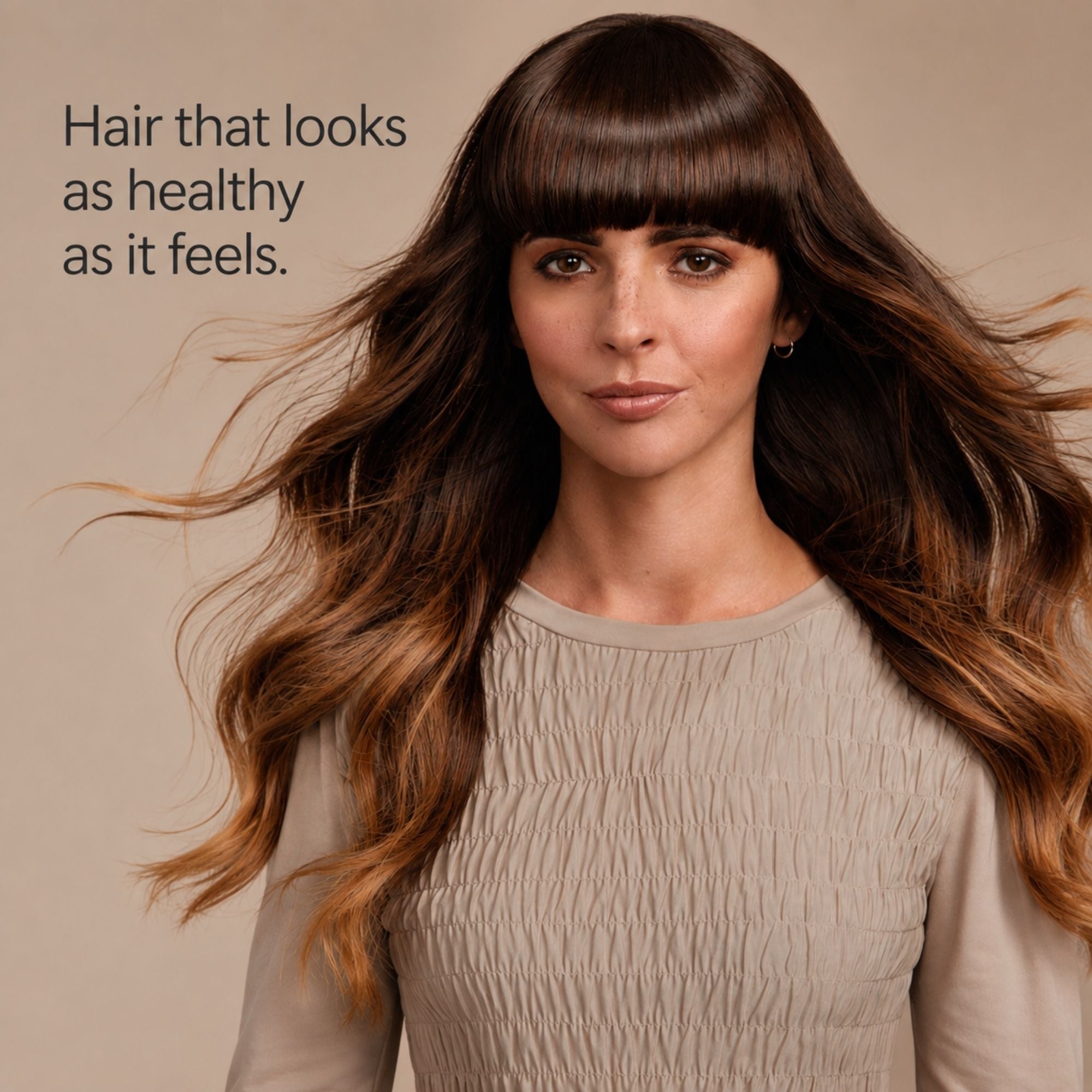 A woman with long, wavy brown hair and straight bangs wearing a beige textured top, set against a solid beige background with the text 'Hair that looks as healthy as it feels.'.