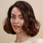 A close-up portrait of a woman with a short, wavy brown wig with subtle warm caramel highlights, set against a solid beige background.