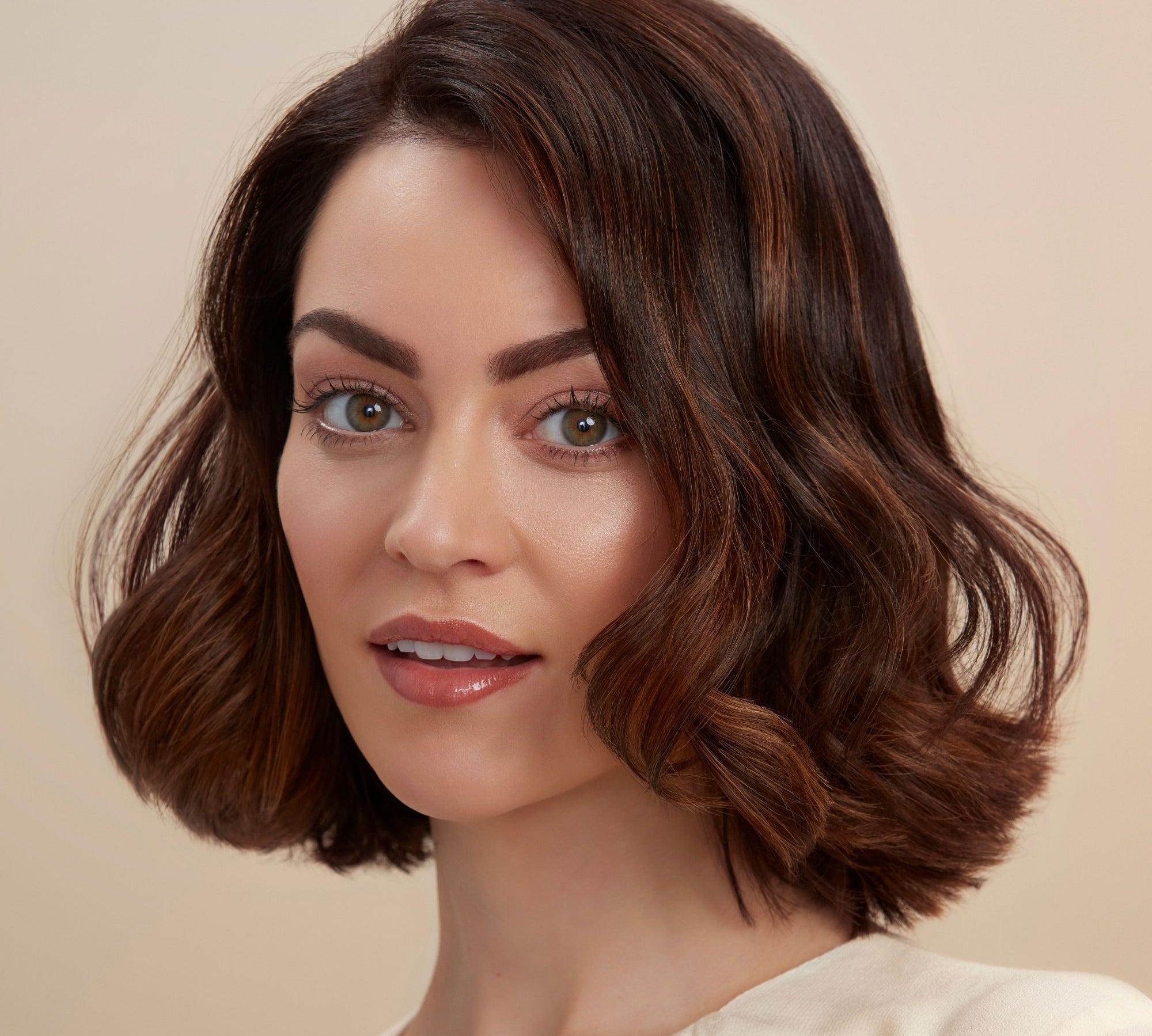 A close-up portrait of a woman with a short, wavy brown wig with subtle warm caramel highlights, set against a solid beige background.