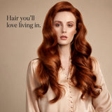 A woman with long, wavy auburn-red hair wearing a light beige satin blouse, posed against a warm beige background with the text 'Hair you'll love living in.' on the left.