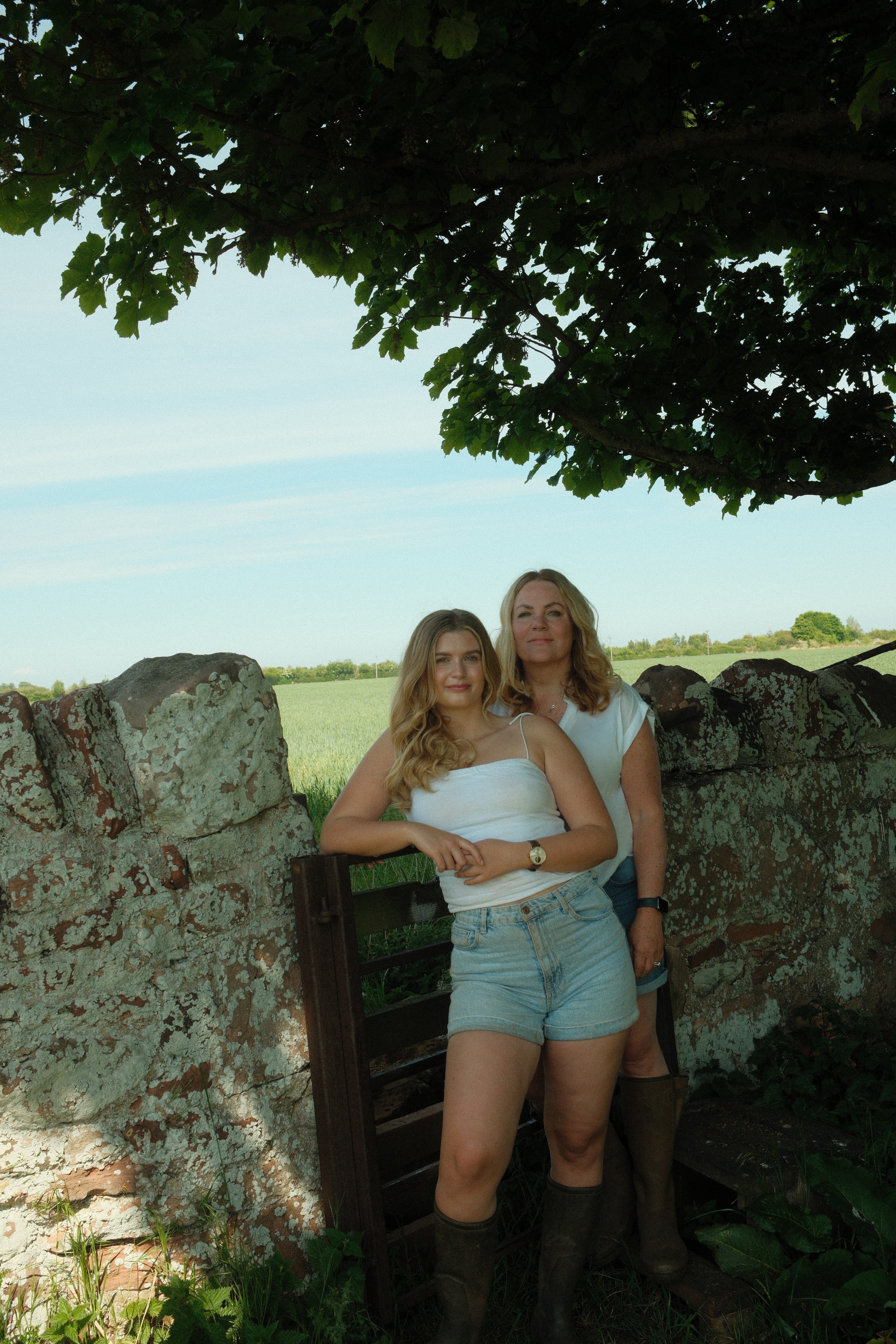 our founder mother daughter duo standing next to a stone wall with a field and blue sky in the background both in white tops and denim shorts with blonde hair