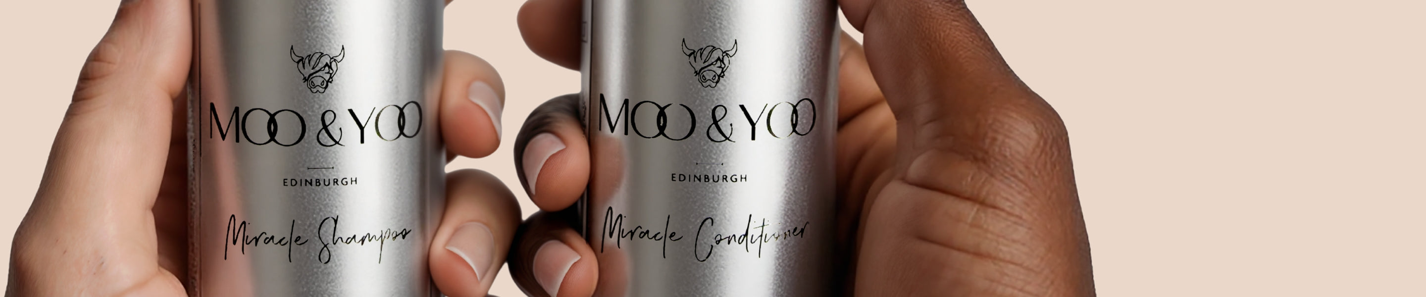 Two bottles of Moo & Yoo shampoo and conditioner in aluminium mini travel bottles held by hands against a beige background