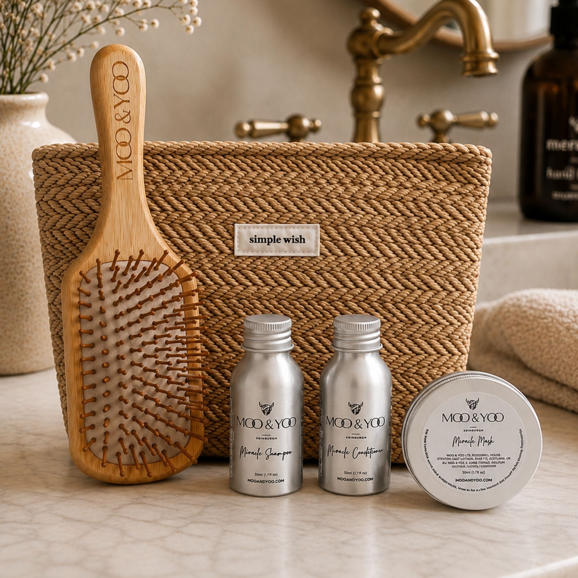 A hair care gift set from MOO & YOO on a marble bathroom counter, featuring a woven toiletries pouch, a wooden paddle hairbrush, two silver bottles of Shampoo and Conditioner, and a round tin of Mask, with a brass tap and vase in the background.