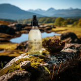 A glass jar of Moo and Yoo Volumising Spray Mist on a rock covered in moss with a small glass bowl of moss to the right. It is overlooking a small loch in Scotland with the hills in the distance.