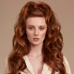 A close-up portrait of a woman with long, voluminous, coppery red wavy hair styled in a high half-up bun with loose curls. She is wearing a cream-colored ribbed button-up shirt against a solid warm beige background.
