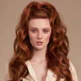 A close-up portrait of a woman with long, voluminous, coppery red wavy hair styled in a high half-up bun with loose curls. She is wearing a cream-colored ribbed button-up shirt against a solid warm beige background.