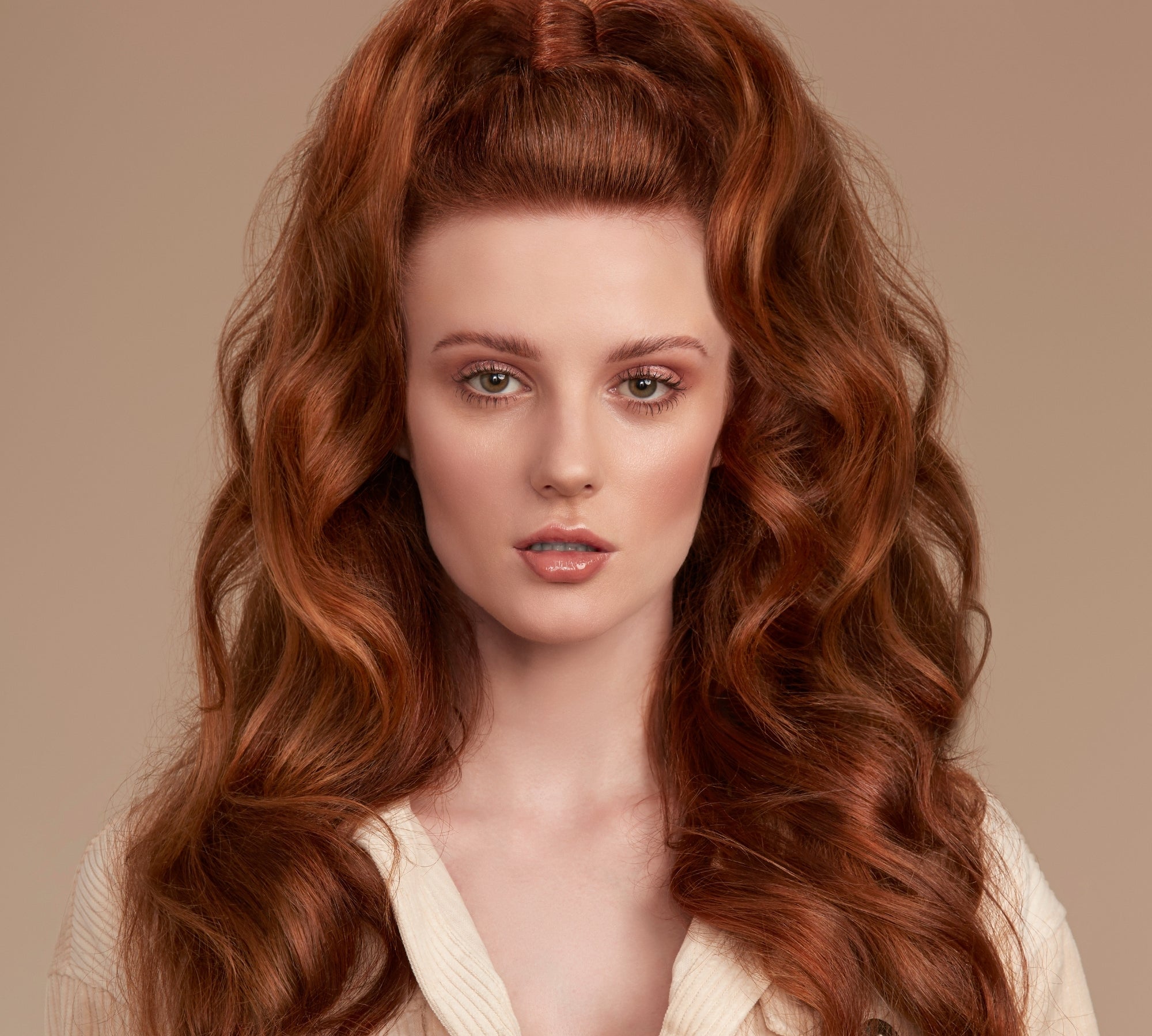 A close-up portrait of a woman with long, voluminous, coppery red wavy hair styled in a high half-up bun with loose curls. She is wearing a cream-colored ribbed button-up shirt against a solid warm beige background.