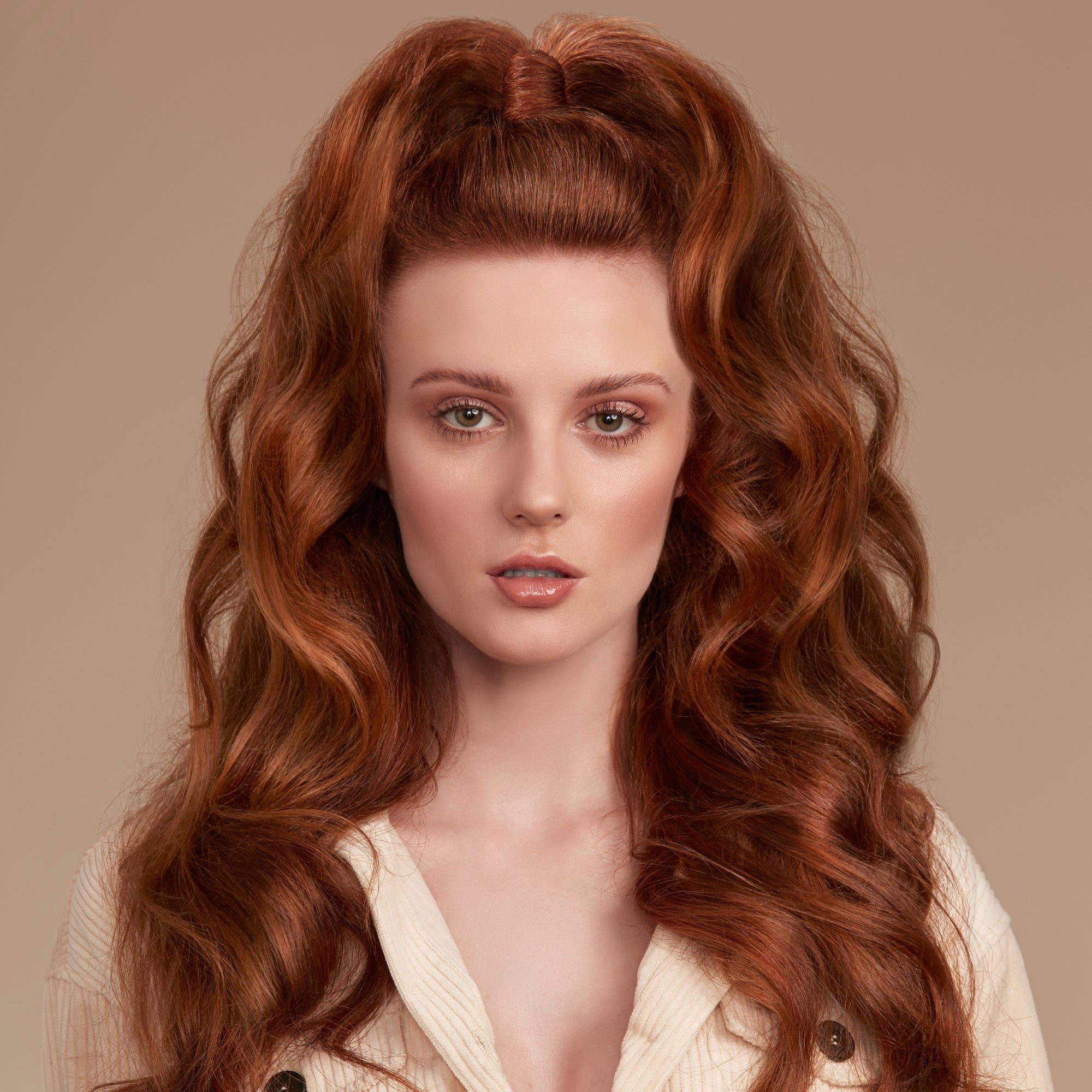 A close-up portrait of a woman with long, voluminous, coppery red wavy hair styled in a high half-up bun with loose curls. She is wearing a cream-colored ribbed button-up shirt against a solid warm beige background.