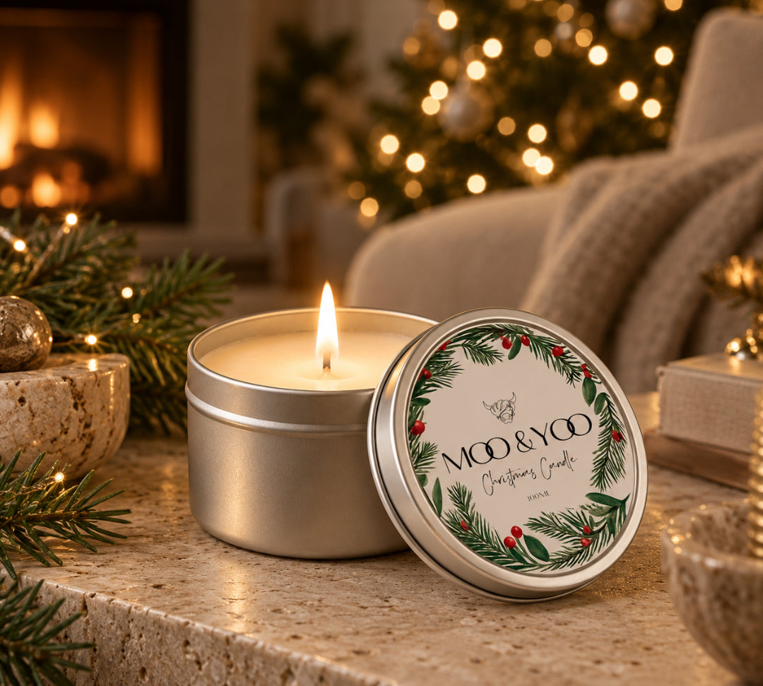 Moo & Yoo Christmas Candle in a silver tin, lit on a natural stone table with festive greenery, a glowing Christmas tree and a cosy fireplace in the background.