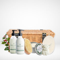Luxury body care product hamper with our vegan body wash and body lotion in glass bottles, a sisal dry body brush, christmas scented tin vegan candle, body buffer pad and a highland cow shaped hand soap all infront of a wicker hamper basket with buckles against a white background 
