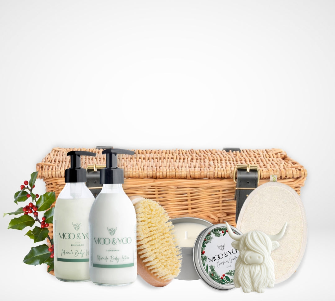 Luxury body care product hamper with our vegan body wash and body lotion in glass bottles, a sisal dry body brush, christmas scented tin vegan candle, body buffer pad and a highland cow shaped hand soap all infront of a wicker hamper basket with buckles against a white background 