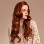 Female model with flowing long red wavy hair from using Moo and Yoo Miracle Conditioner. She has a pale cream lacey top on.  She is looking to the right so you can see the full side of her hair. She has super shiny long wavy hair using Moo and Yoo conditioner with lots of volume.