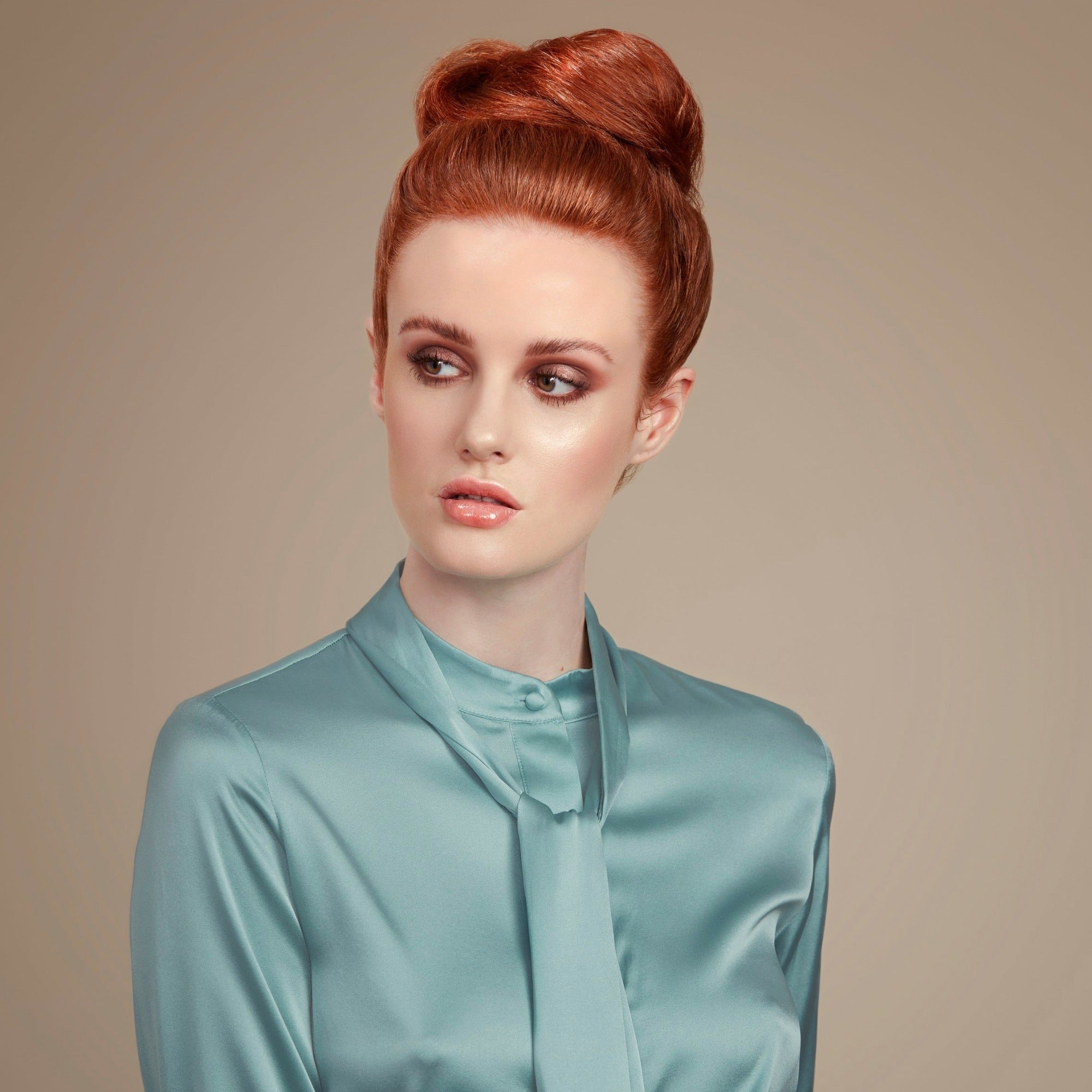 Female model with red hair in a high bun held in place with Moo and Yoo hair spray.  She is facing the camera and looking to her left. She is wearing a pale blue top that is long sleeved and has a high neck. 