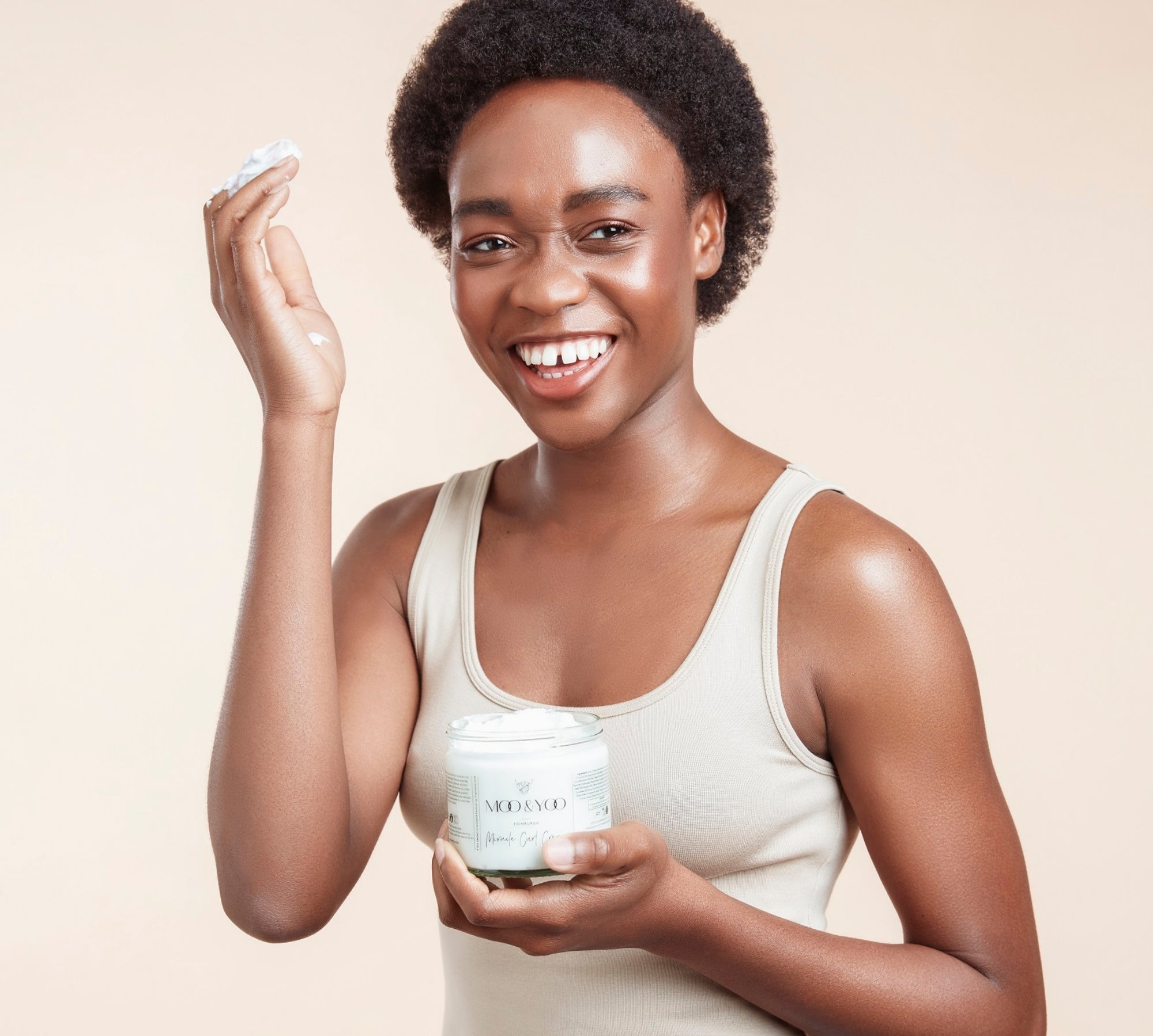 Woman with cropped afro hair holding a jar of Moo & Yoo Miracle Curl Cream in one hand and her other hand up to her hair with product on it with a beige background