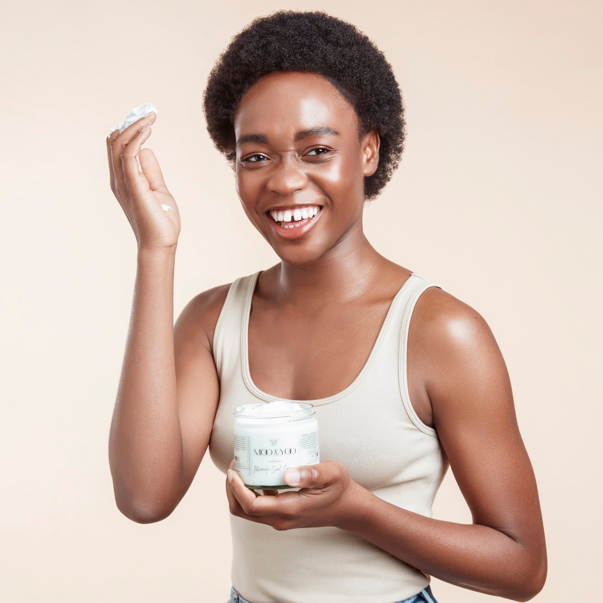 Woman with cropped afro hair holding a jar of Moo & Yoo Miracle Curl Cream in one hand and her other hand up to her hair with product on it with a beige background