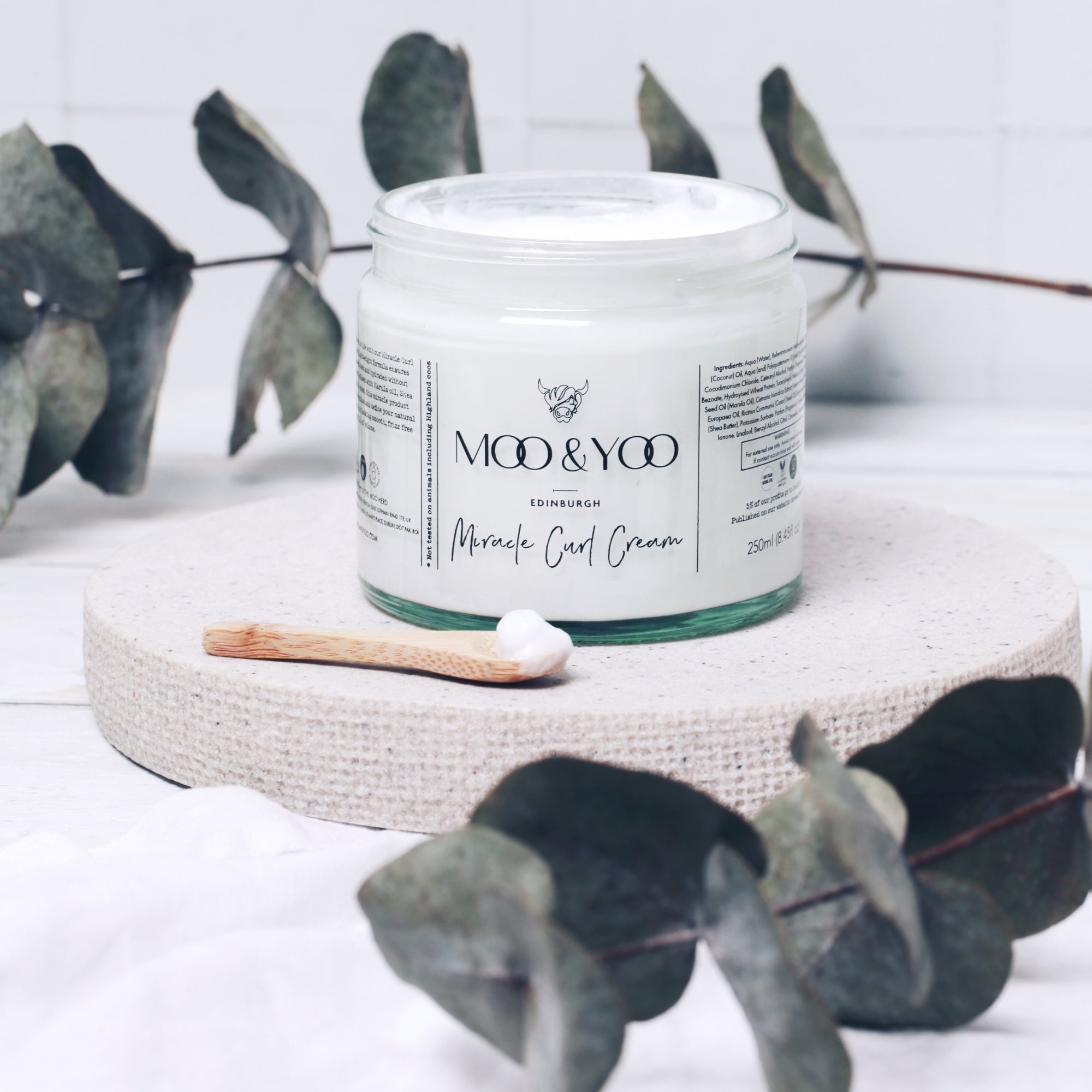 Jar of 'Moo & Yoo' curl cream on a textured surface with eucalyptus leaves in the background