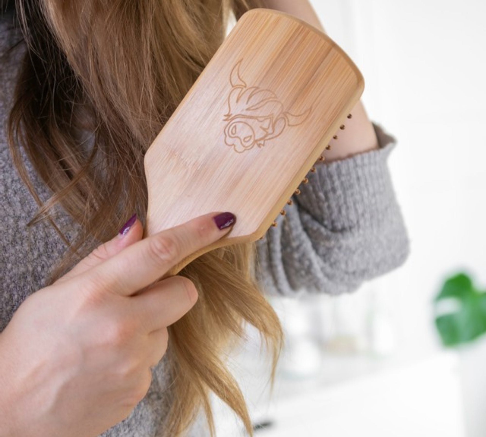 Person brushing their blond hair with wooden hairbrush with Moo & Yoo cow logo, wearing grey jumper