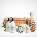 Hamper of hair care products including glass shampoo and conditioner bottles, a vegan christmas candle, a bamboo hair brush, and a small cow shaped hand soap, a fabric eco net shower ramie and a wicker hamper with black straps and buckles on a white background.