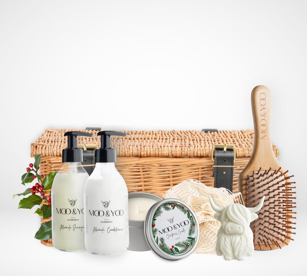 Hamper of hair care products including glass shampoo and conditioner bottles, a vegan christmas candle, a bamboo hair brush, and a small cow shaped hand soap, a fabric eco net shower ramie and a wicker hamper with black straps and buckles on a white background.